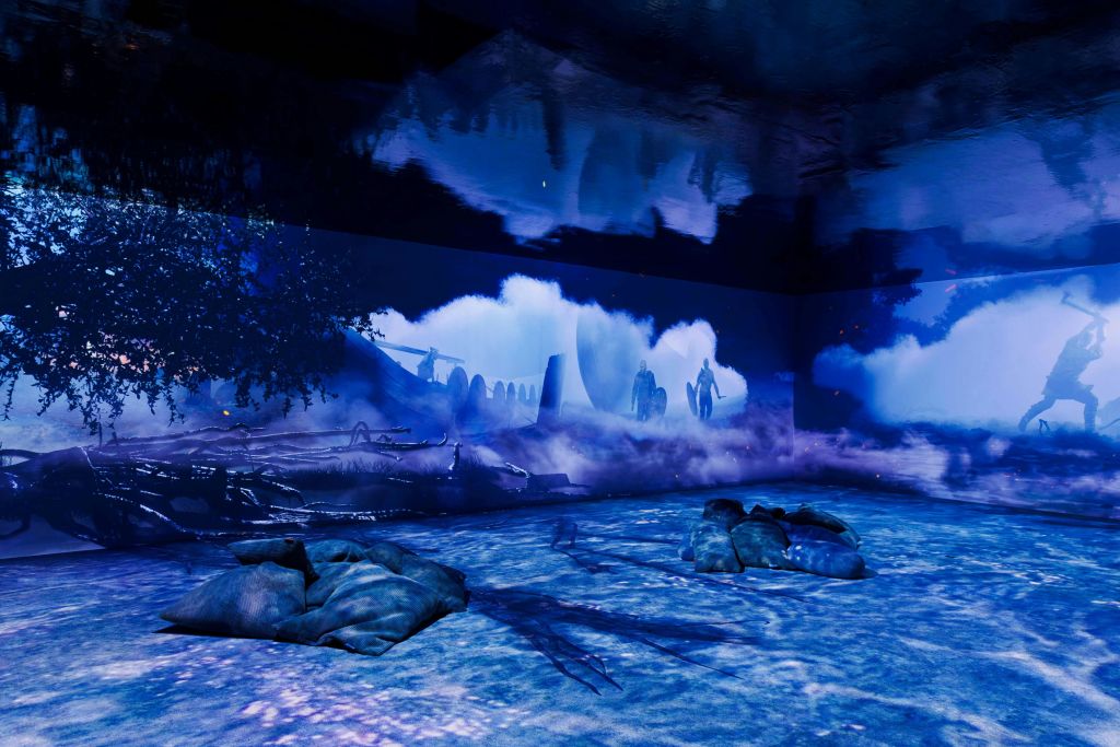 파란색으로 빛나는 방으로, 사람들의 실루엣과 거대한 달이 있는 신비로운 풍경의 벽화가 특징입니다. 베개가 바닥에 흩어져 있습니다.