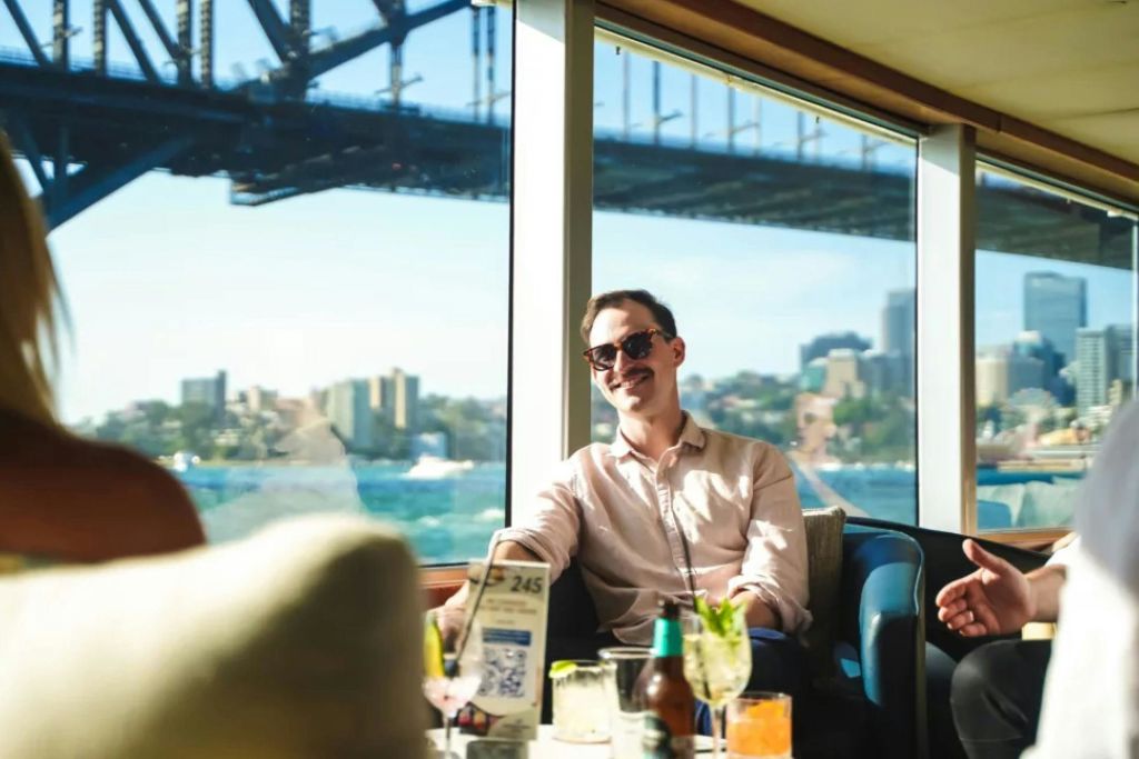 창문 너머로 도시 풍경과 다리가 보이는 라운지 공간에 선글라스를 착용한 남성이 앉아 있습니다.