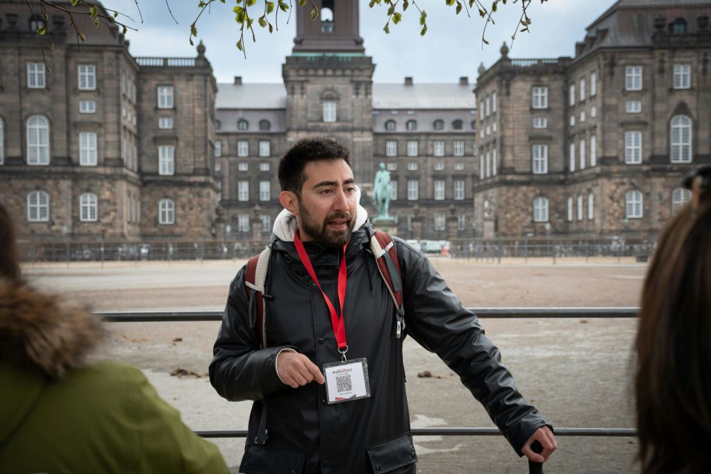 커다란 역사적인 건물 앞에서 방아끈을 두른 남성이 말을 건네고 있으며, 앞쪽에 또 다른 남성이 부분적으로 보입니다.