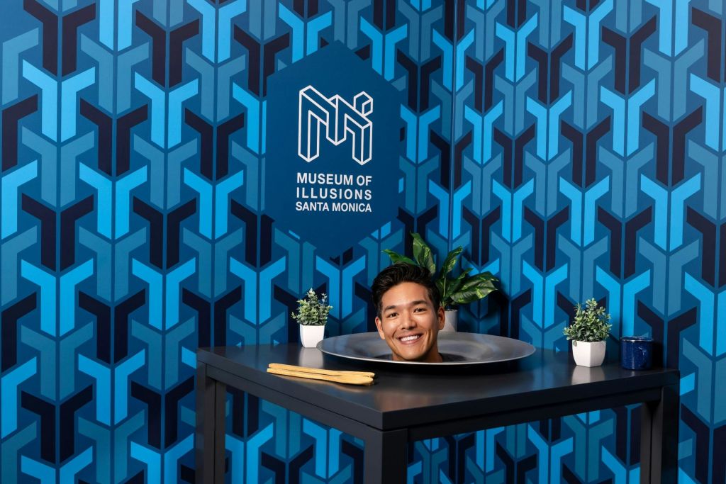한 사람이 테이블 위에 놓인 접시에 기하학적 파란색 배경의 산타모니카 환상 박물관 (Museum of Illusions Santa Monica) 이라는 라벨이 붙은 플래터에 머리를 대고 있는 것으로 보입니다.