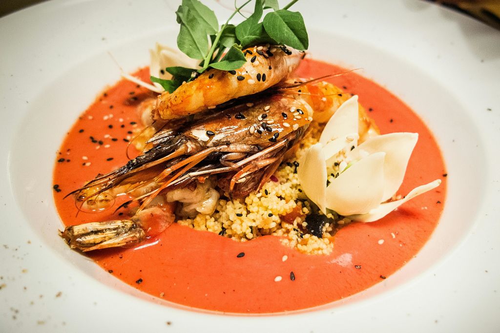 녹색과 씨앗으로 장식된 쿠스쿠스 침대에 새우를 올려 놓은 미식가 요리입니다. 붉은 소스에 하얀 야채 조각이 들어 있습니다.