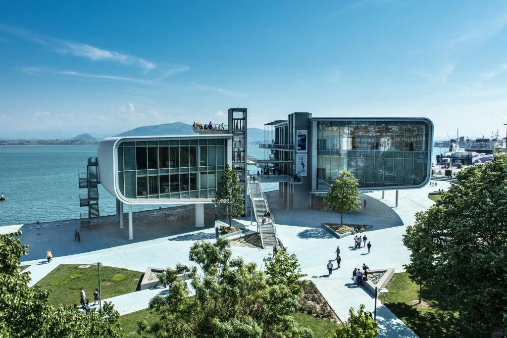 유리 파사드가 있는 현대적인 수변 건물, 높은 산책로, 야외 계단. 걷는 사람들과 흩어져 있는 나무들.