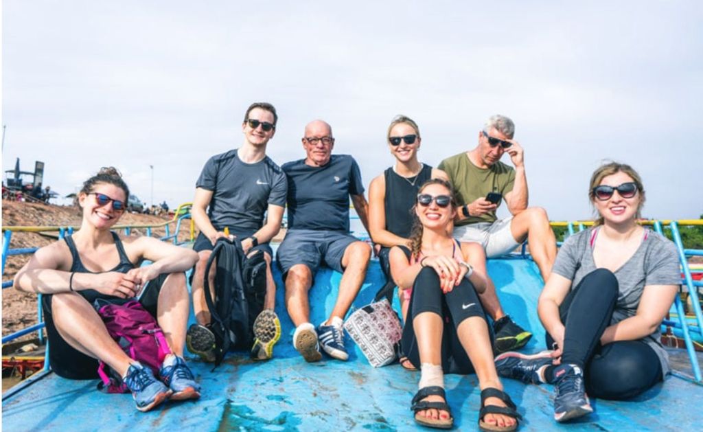 여섯 명이 야외에서 파란색 표면에 앉아 웃으며 캐주얼한 옷과 선글라스를 착용하고 있습니다.