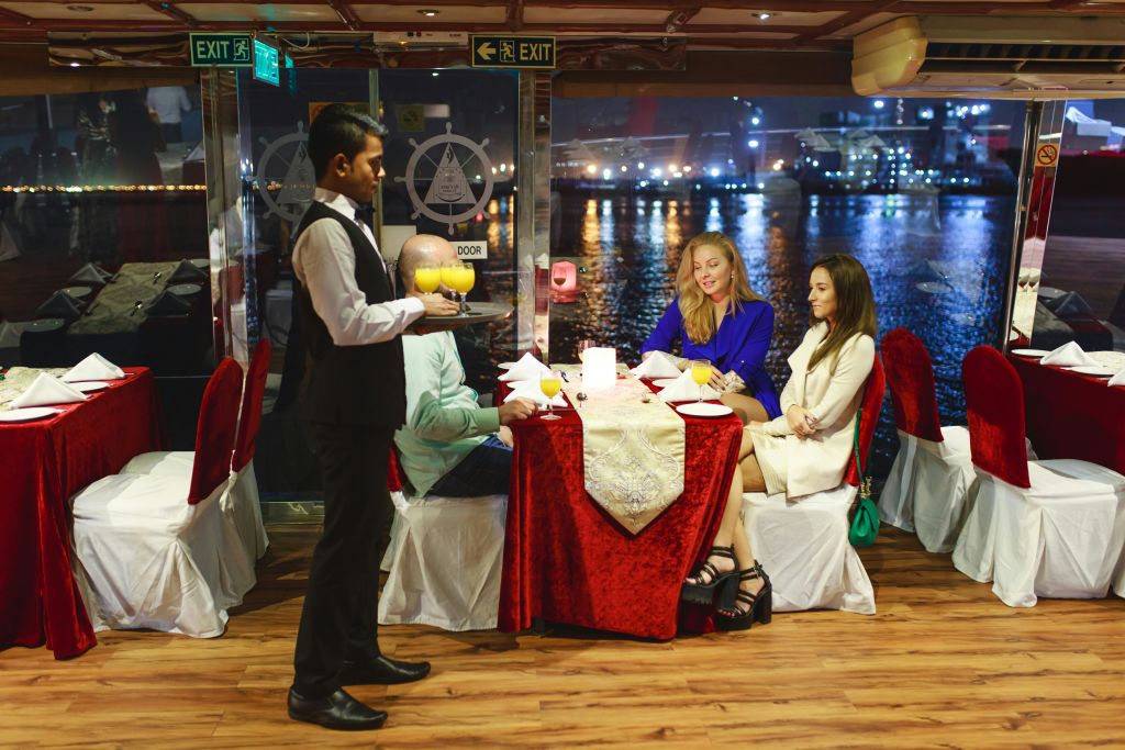 웨이터가 빨간색 식탁보가 놓인 테이블에서 앉아 있는 세 명의 손님에게 음료를 제공합니다. 물에 반사되는 도시의 불빛을 배경으로 합니다.