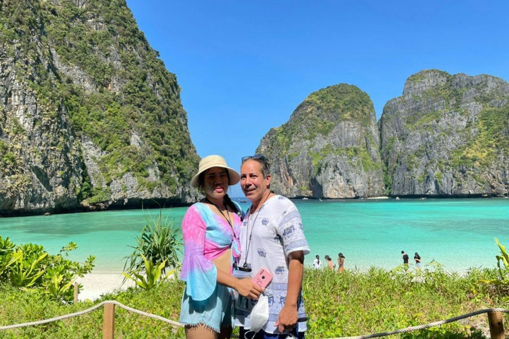 청록색의 맑은 물과 푸른 하늘을 배경으로 커다란 바위 절벽이 펼쳐진 열대 해변 앞에 커플이 서 있습니다.