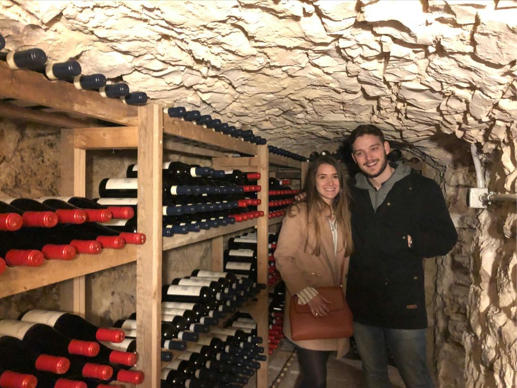 와인 병이 가득한 선반으로 둘러싸인 와인 저장고에 서 있는 두 사람.