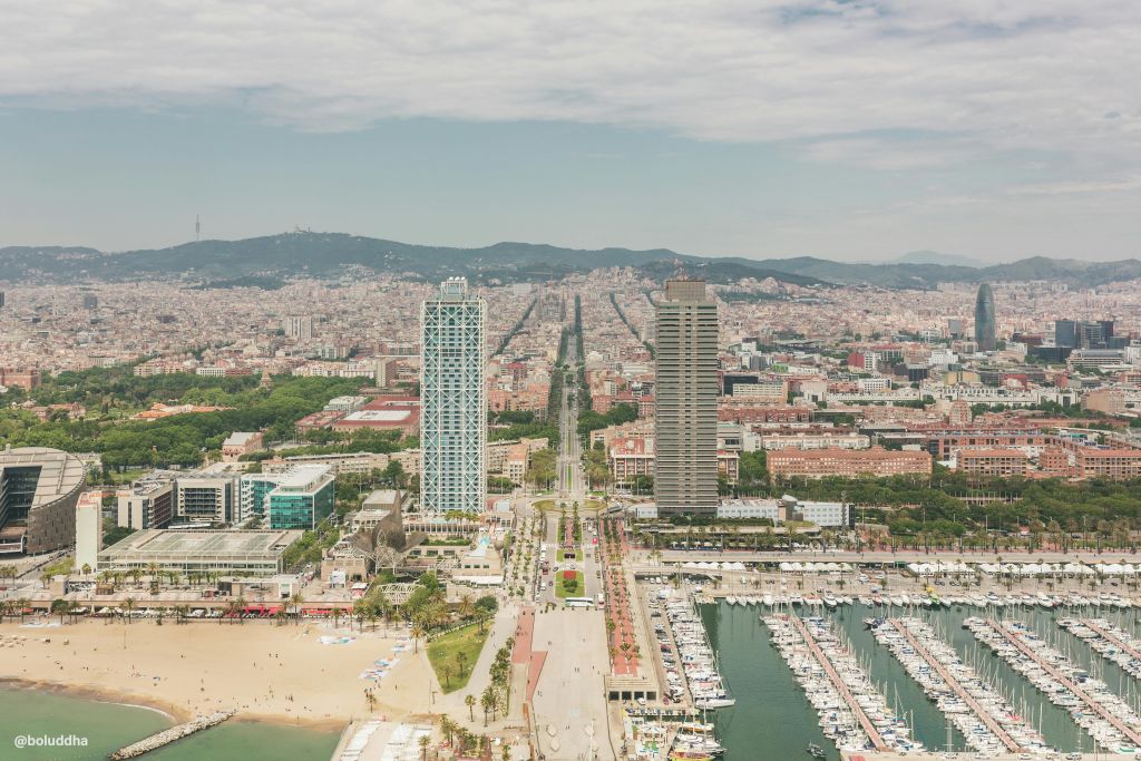 두 개의 높은 건물, 보트가 있는 정박지, 사람들이 있는 모래 해변이 있는 바르셀로나 해안선의 공중 전망을 감상할 수 있습니다.