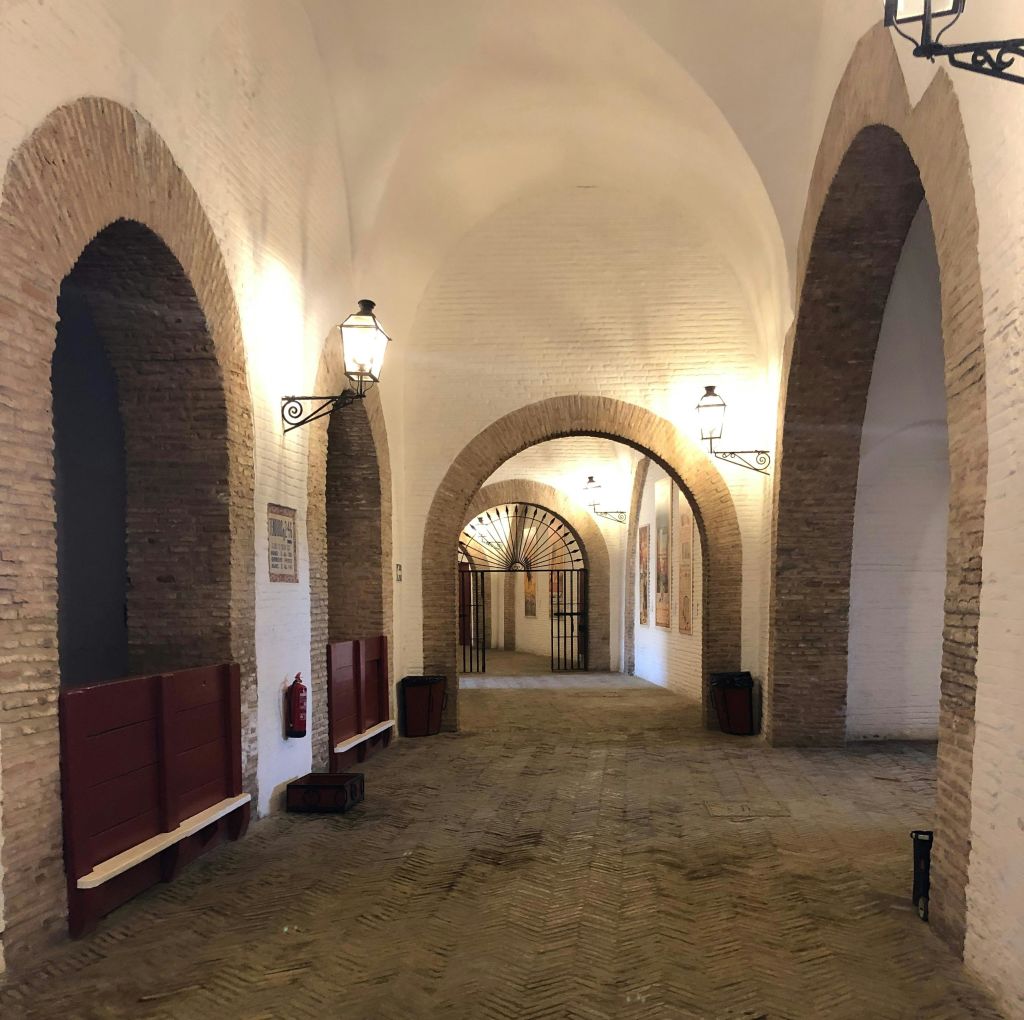 아치형 벽돌로 된 출입구, 벽 조명, 벤치, 소화기, 금속 게이트가 있는 복도.