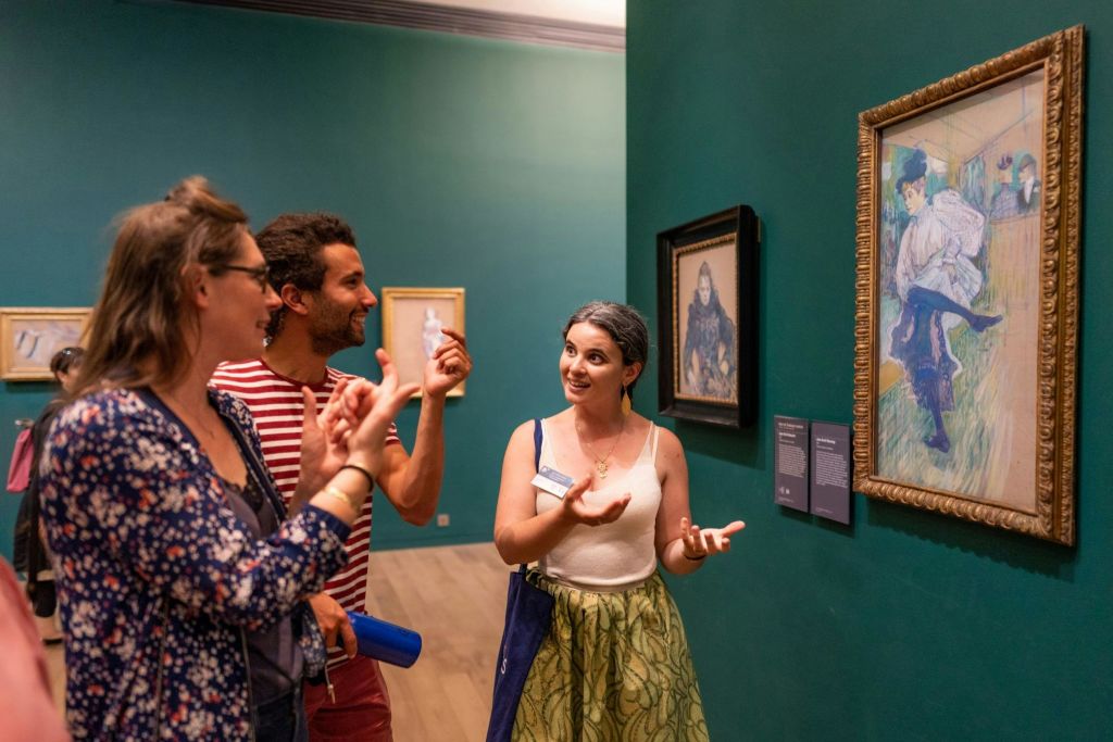 벽에 그림이 있고 청록색 배경이 있는 미술관에서 세 사람이 대화를 나누고 있습니다.