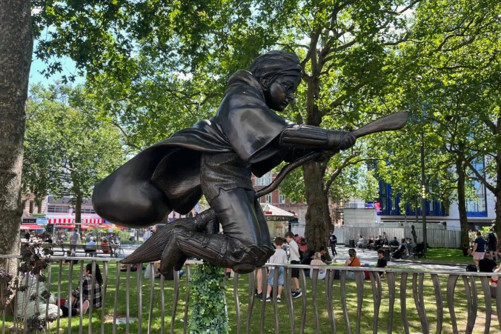 나무와 사람을 배경으로 공원을 배경으로 빗자루를 타고 날아다니는 사람의 청동 동상.