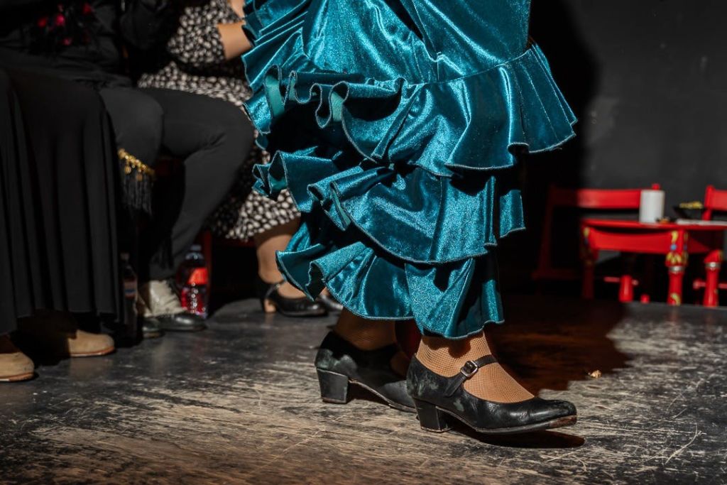청록색 러플 드레스와 검은색 굽이 달린 신발을 신고 있는 사람이 나무 바닥에서 춤을 추고 있으며, 좌석에 앉아 관람객을 배경으로 춤을 추고 있습니다.