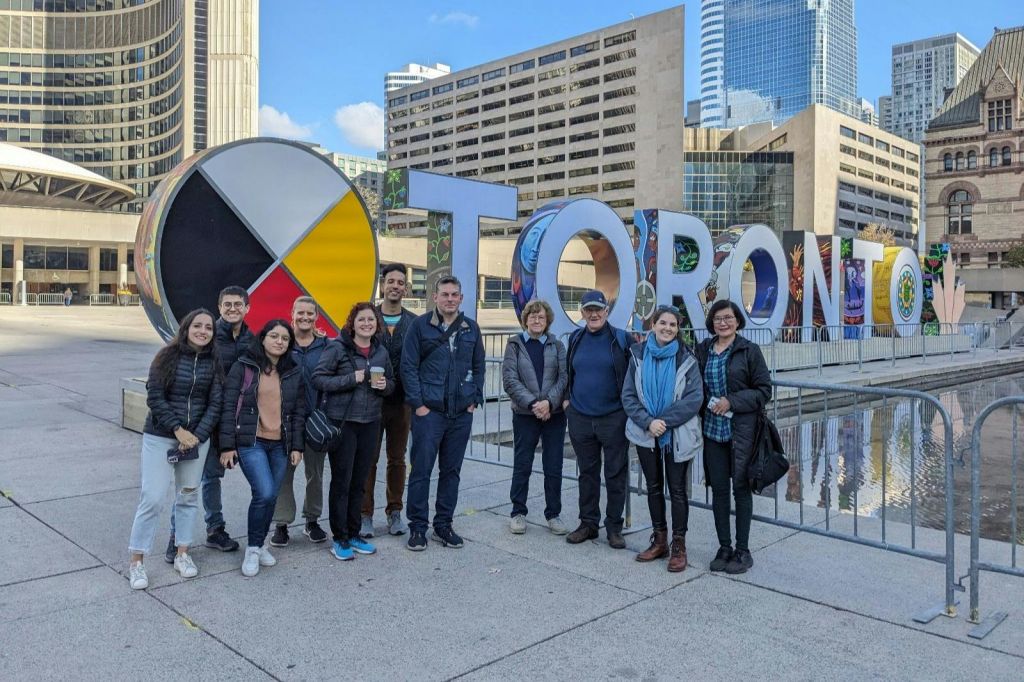 현대적인 건물이 있는 도심 광장의 화려한 글자가 적힌 커다란 간판 앞에 한 무리의 사람들이 서 있습니다.