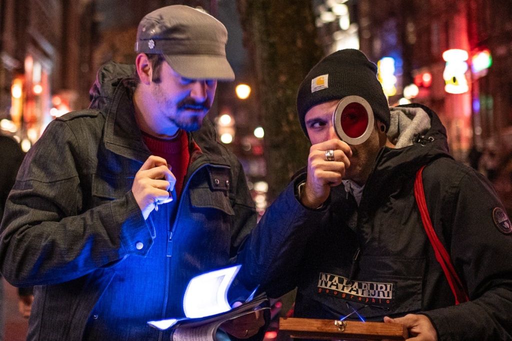 밤에 책 한 권과 빨간색 렌즈를 살펴보는 두 남자의 모습이 푸른 빛에 비춰졌고, 도시의 불빛이 배경으로 흐릿하게 비춰졌습니다.