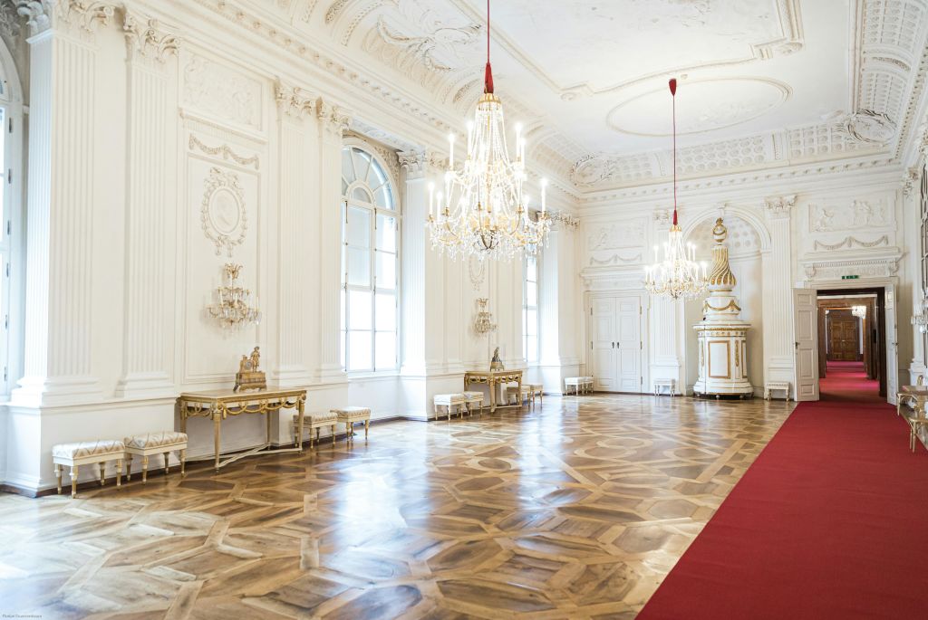 샹들리에, 쪽모이 세공 마루, 흰색 벽, 금색 가구, 레드 카펫이 깔린 통로가 있는 넓고 화려한 객실입니다.