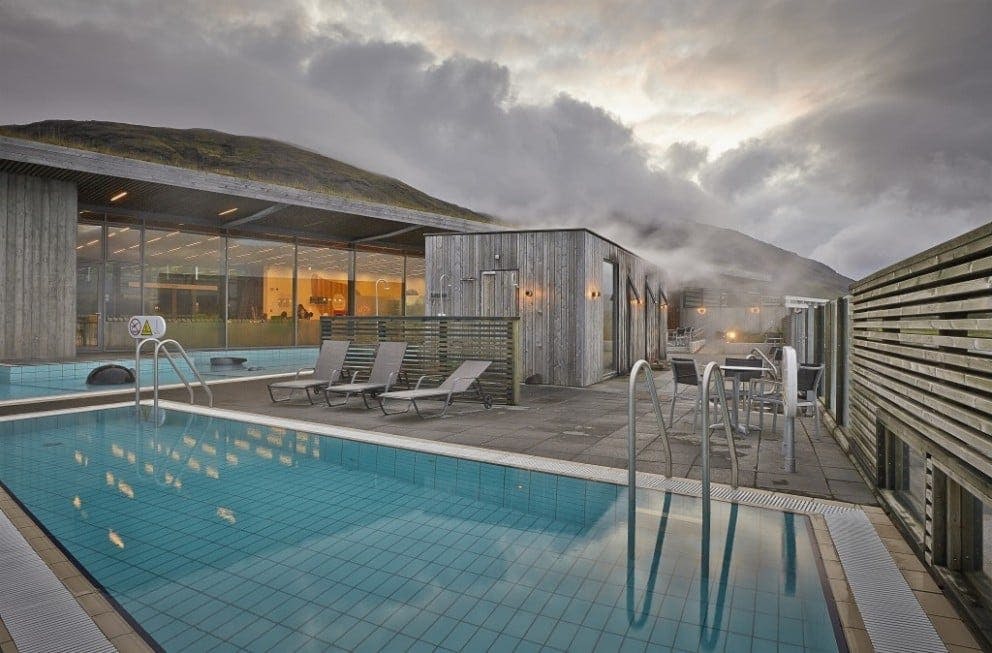 안락의자, 목조 건물, 수증기가 떠오르는 현대적인 야외 수영장 공간. 산과 흐린 하늘을 배경으로