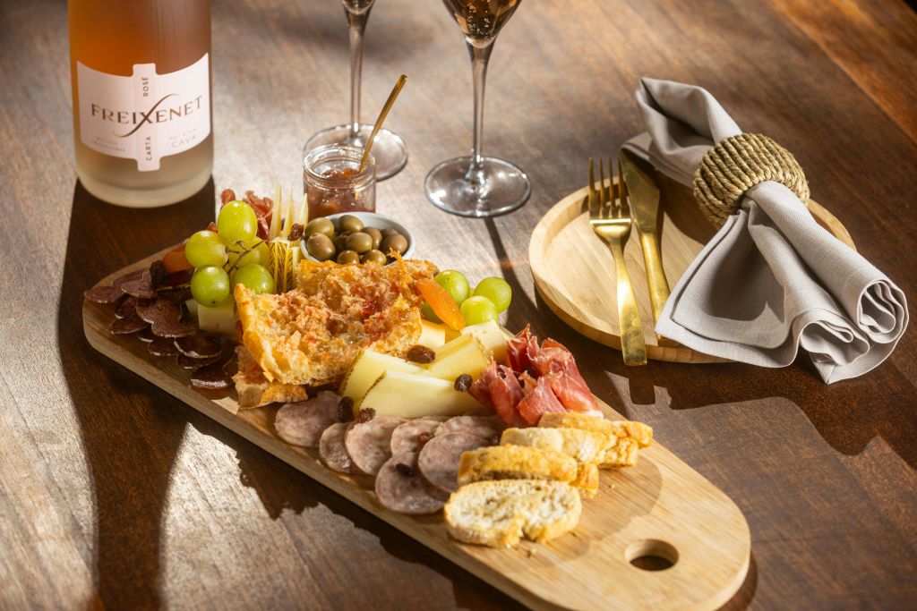 다양한 치즈, 고기, 포도, 올리브, 빵, 잼이 담긴 샤퀴트리 보드. 근처에 와인잔, 수저, 냅킨이 있습니다.