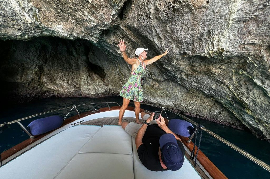 한 여성이 바위 동굴 아래 보트에 서서 한 남성이 카메라를 들고 누워 있는 동안 지붕을 잡고 있습니다.
