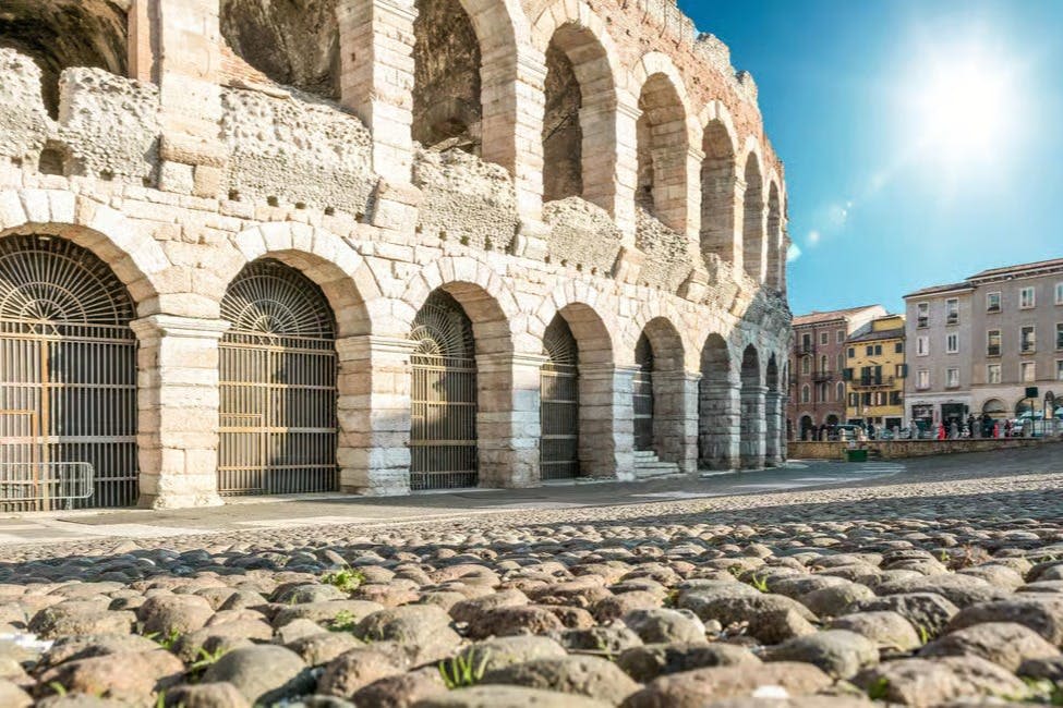 맑고 푸른 하늘 아래 아치형 문과 자갈 전경이 있는 고대 석조 원형극장. 주변 건물이 부분적으로 보입니다.