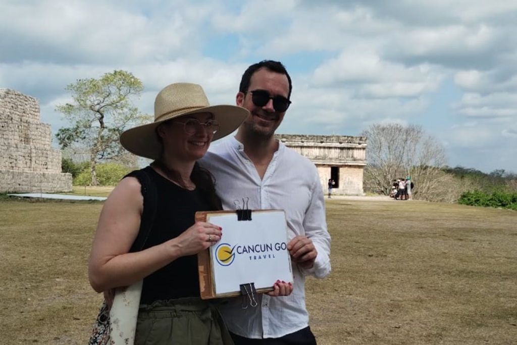 두 사람이 '칸쿤 고 트래블 (Cancun Go Travel)' 이라는 팻말을 들고 열린 공간에 서서 미소를 짓고 있습니다. 배경에는 유적과 나무가 보입니다.