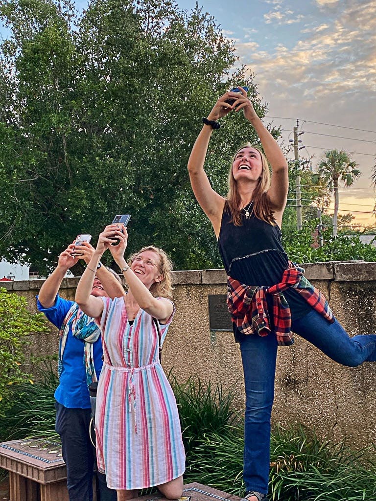 나무와 흐린 하늘을 배경으로 세 사람이 열정적으로 휴대폰을 들고 미소를 짓고 있습니다.