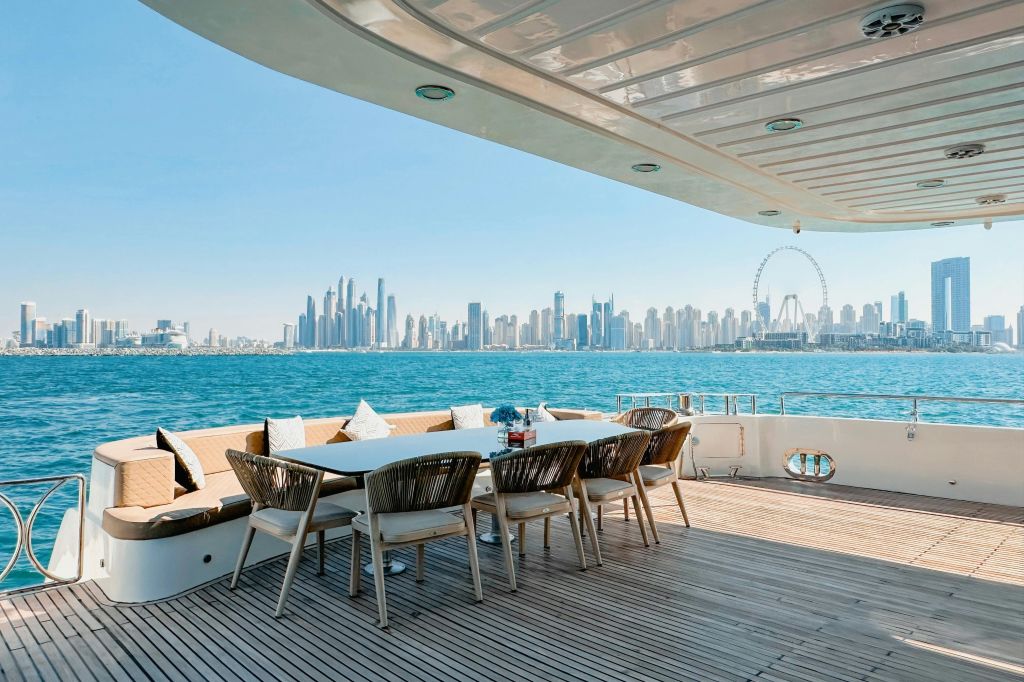 고리 버들 의자와 테이블이 있는 요트의 야외 식사 공간으로, 도시의 스카이라인과 물 건너편에 있는 커다란 전망대가 내려다보입니다.