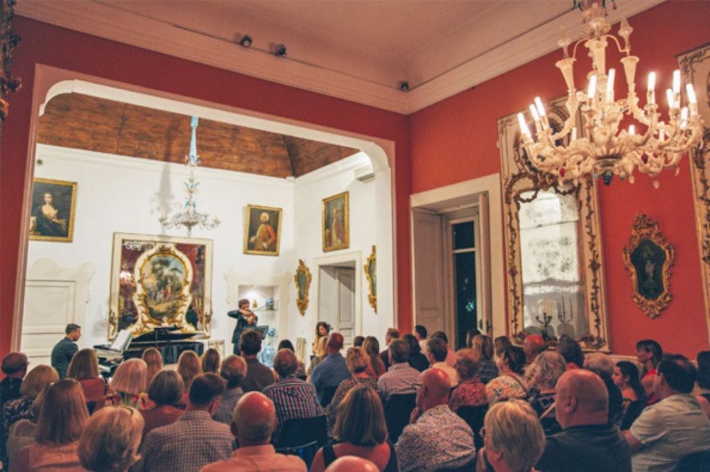 그림과 샹들리에가 있는 정교하게 꾸며진 방에서 한 여성이 청중 앞에서 바이올린을 타고 공연하고 있습니다.