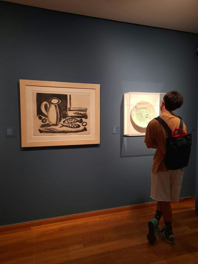 파란색 박물관 벽에 두 개의 액자 작품을 관찰하는 배낭이 있는 사람. 하나는 흑백 드로잉이고 다른 하나는 원형 조각입니다.