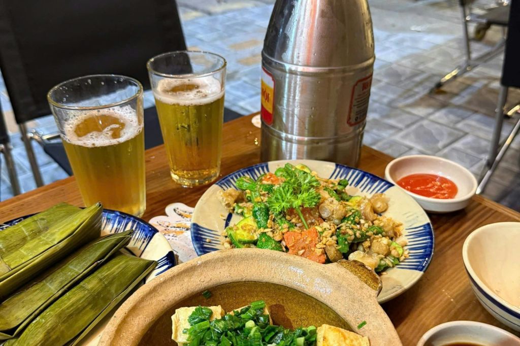 맥주 두 잔, 혼합 볶음 접시, 두부 수프 냄비, 포장된 잎 소포, 금속 음료 용기가 있는 테이블.