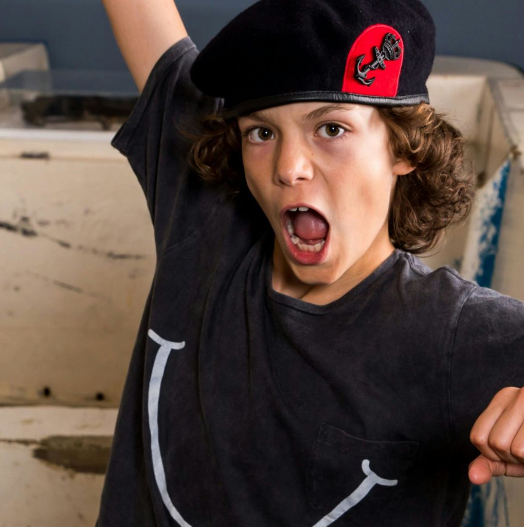 검은 베레모와 회색 티셔츠를 입은 곱슬머리의 젊은이가 실내에서 입을 벌리고 외치고 있습니다.