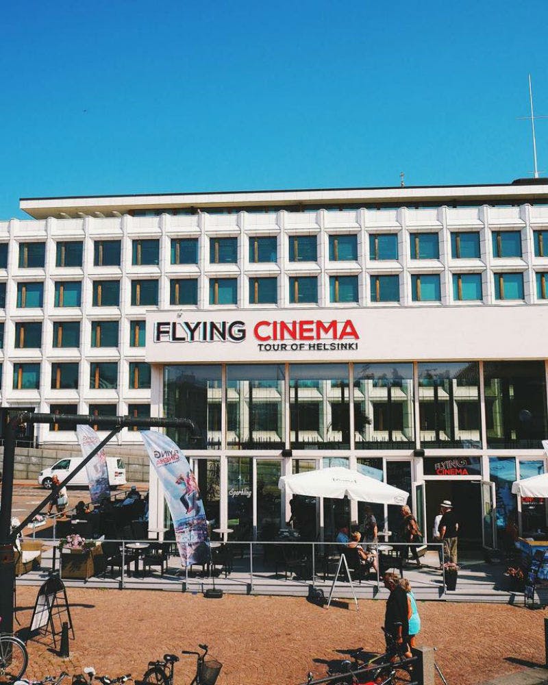 “Flying Cinema Tour Of Helsinki” 표지판, 야외 좌석 공간, 흰색 우산, 몇 명이 있는 현대적인 건물 외관.