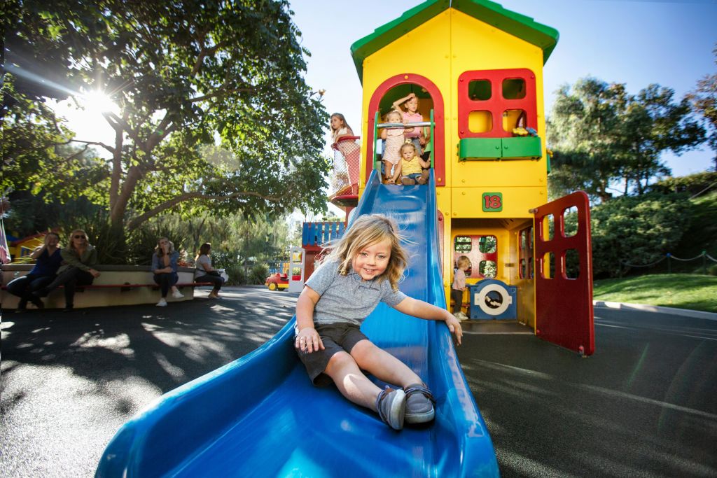 한 어린이가 여러 색상의 놀이방에 부착된 파란색 슬라이드를 아래로 미끄러지듯 내리고, 다른 어린이는 구조물을 배경으로 놀고 있습니다.