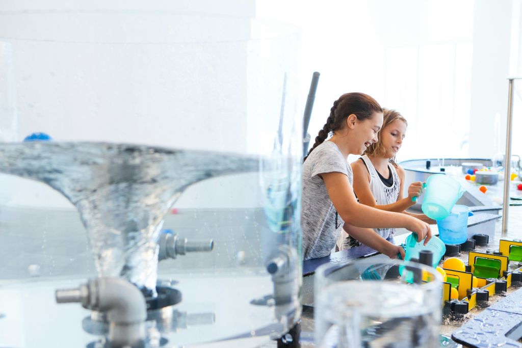 두 어린이가 과학 센터에서 컵, 파이프, 용기를 사용하여 밝고 인터랙티브한 전시물에서 물놀이를 하고 있습니다.