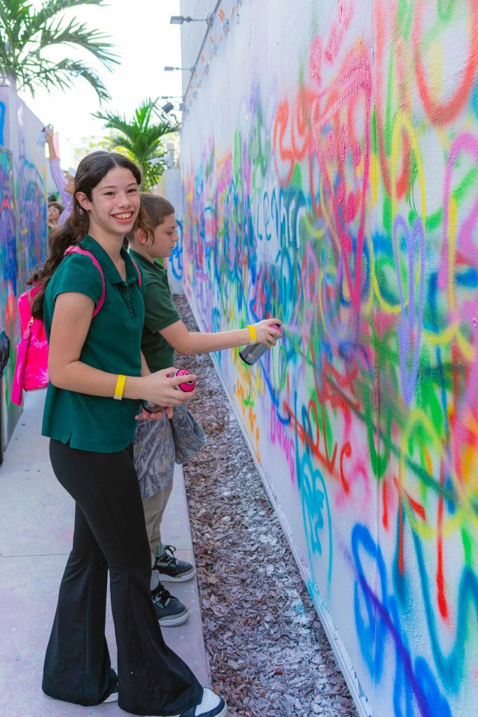 두 명의 소녀가 야외에서 다채로운 색상의 그래피티로 덮인 벽을 그려 웃으며 액티비티에 참여합니다.