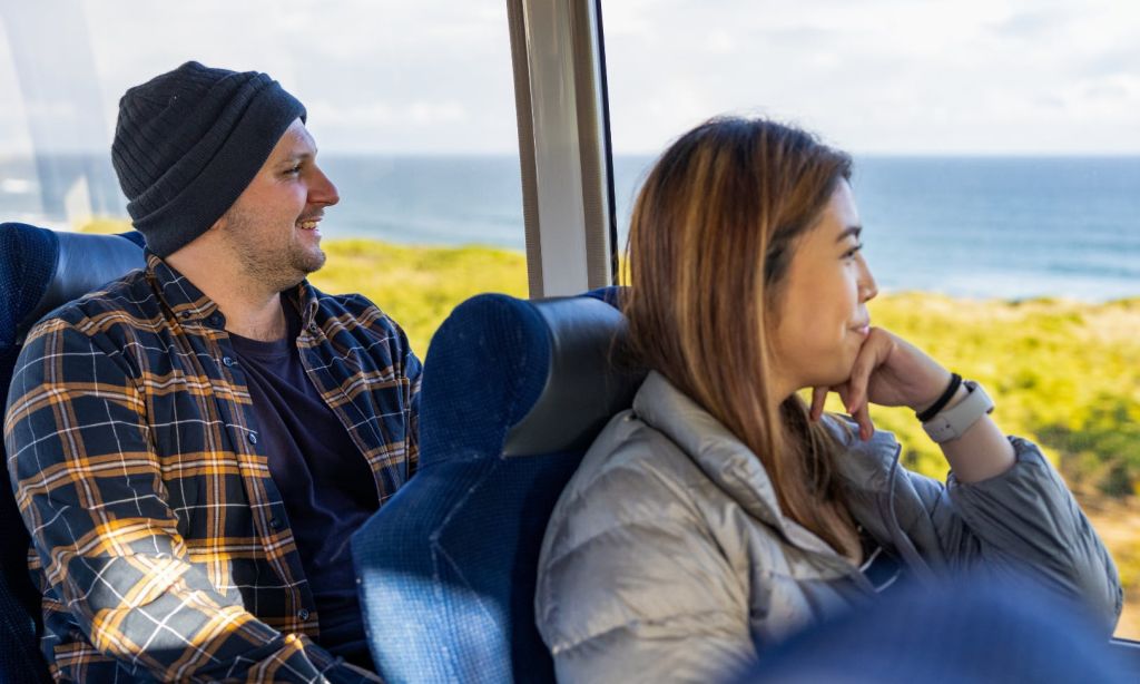 한 남자와 한 여자가 버스에 앉아 녹음과 바다가 어우러진 경치 좋은 풍경을 보며 미소를 짓고 창밖을 내다보고 있습니다.