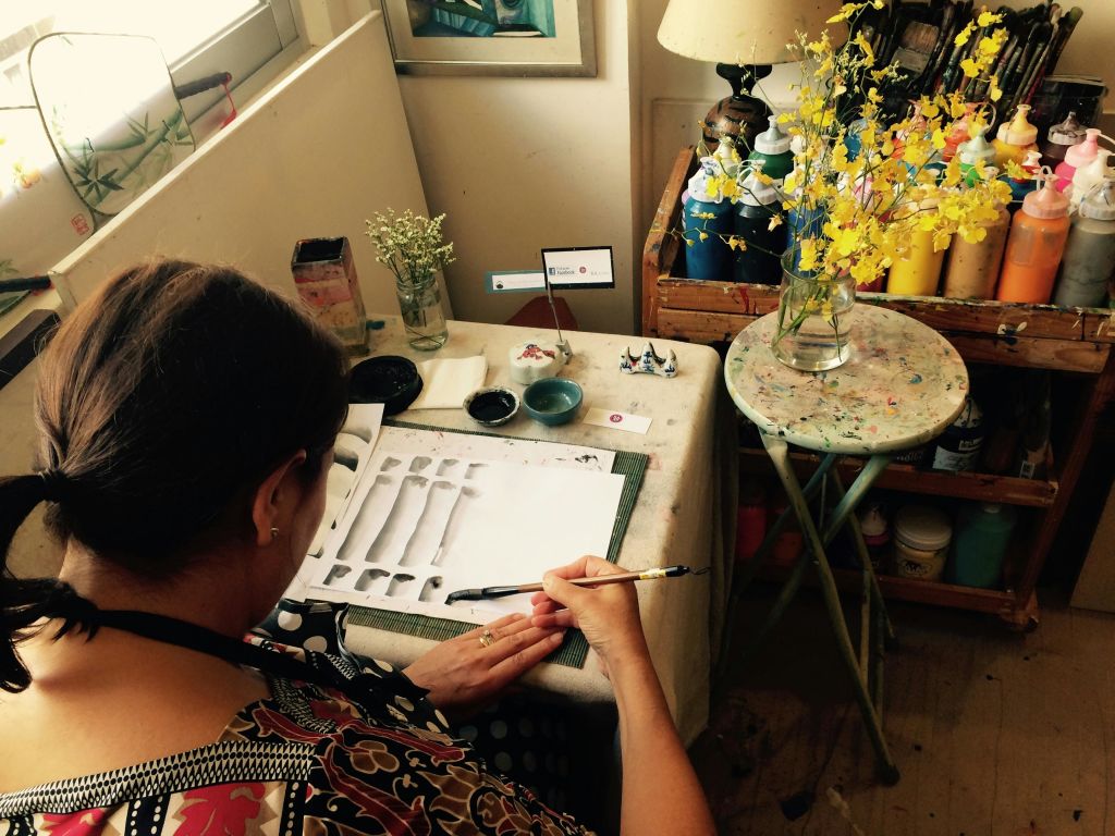 다양한 미술 용품이 놓인 책상, 꽃병에 담긴 꽃, 램프가 놓인 테이블 등 다양한 소모품을 가까이에 두고 그림을 그리는 사람.