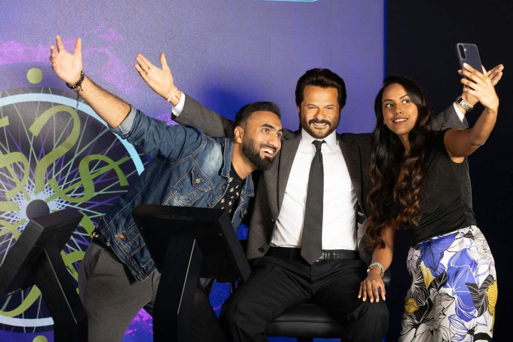 세 사람이 미소를 지으며 셀카를 찍기 위해 포즈를 취하고 있습니다. 양복을 입은 한 남자가 가운데에 앉아 있고, 다른 남자와 한 여자가 옆에 서 있습니다.