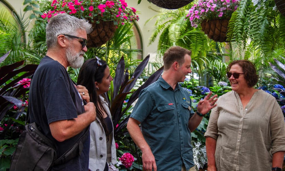 파란색 셔츠와 베이지색 상의를 입은 두 사람이 식물과 꽃이 걸린 울창한 정원에서 웃으며 이야기하고 있습니다.