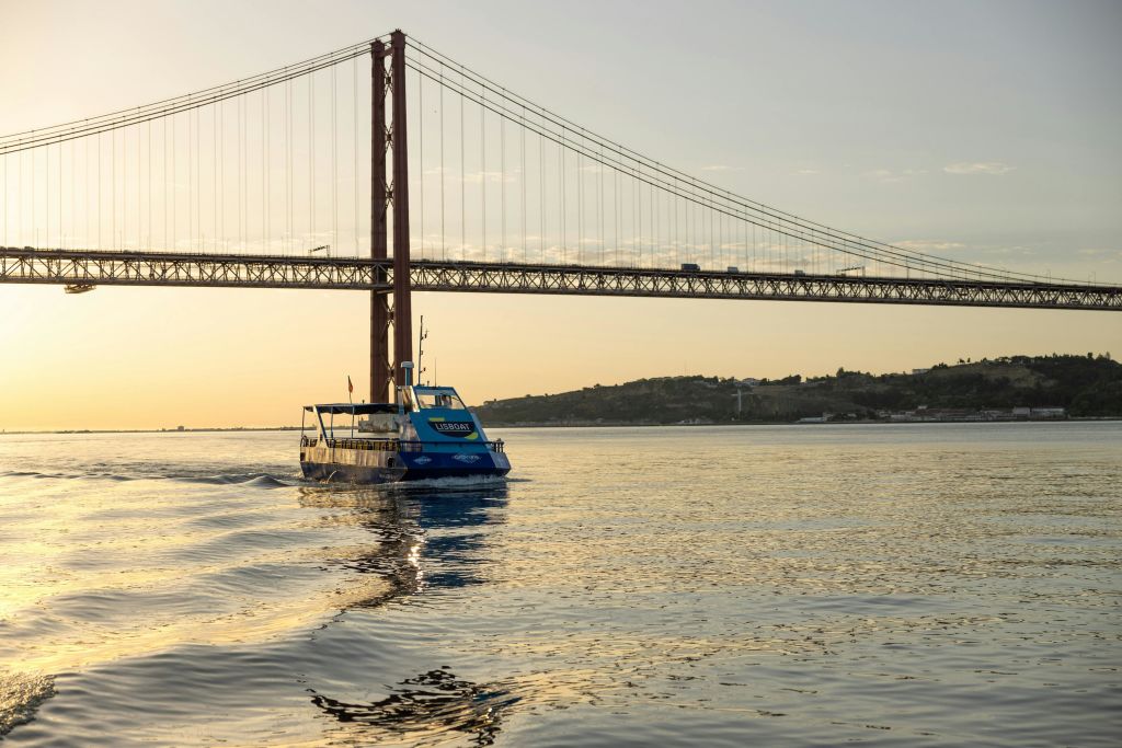 석양을 배경으로 커다란 현수교와 언덕이 있는 잔잔한 바다를 유람하는 푸른 보트입니다.