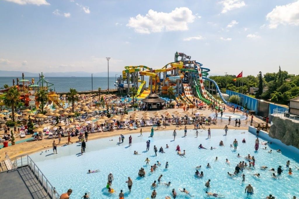 형형색색의 미끄럼틀이 있는 대형 워터파크, 사람들로 붐비는 수영장, 화창한 하늘을 배경으로.