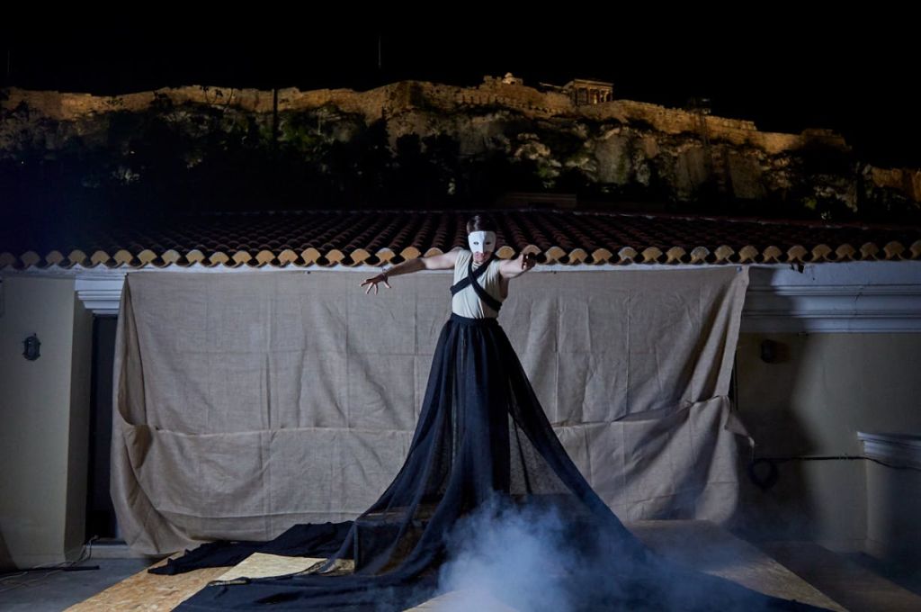 흰색 마스크와 검은색 드레스를 입은 공연자가 무대에서 연기가 자욱한 무대를 배경으로 언덕 꼭대기에 고대의 조명이 켜진 구조물을 배경으로 서 있습니다.