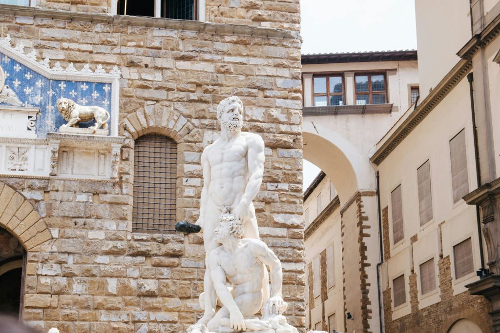 한 명은 서 있고 다른 한 명은 무릎을 꿇은 두 명의 근육질 남성의 대리석 조각상이 아치형 창문이 있는 돌 건물에 세워져 있습니다.