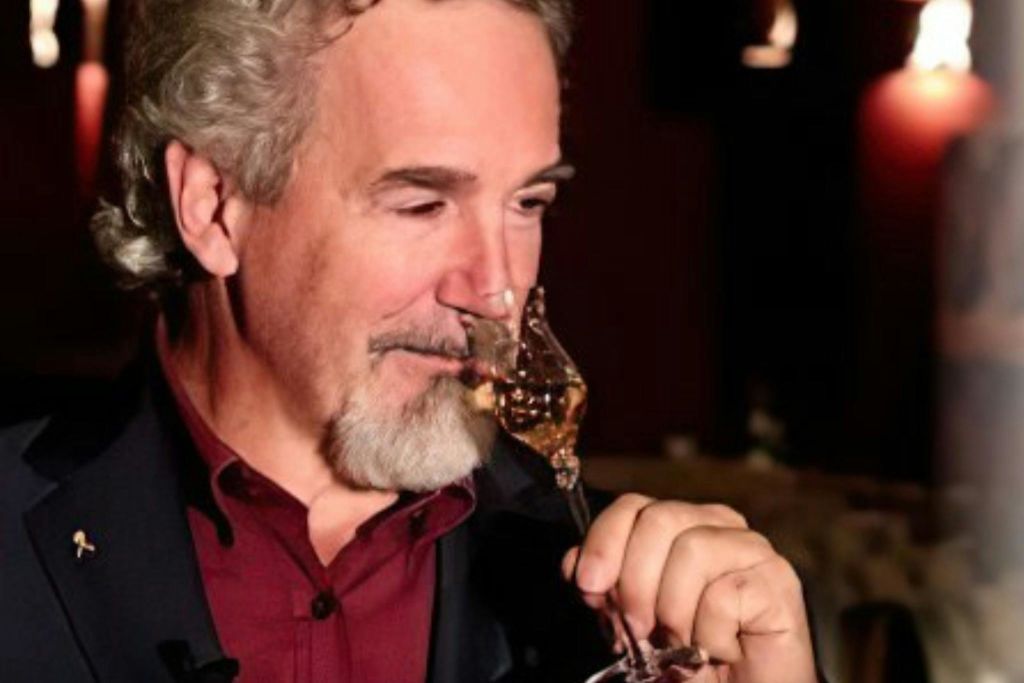 회색 머리카락과 수염을 가진 남자가 어두운 블레이저와 빨간색 셔츠를 입고 희미하게 빛나는 배경에서 호박색 액체를 마시고 있습니다.