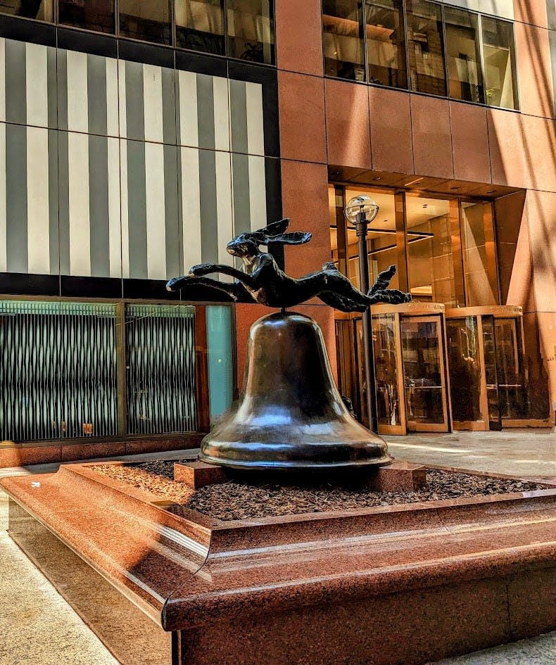 유리와 석조 건물이 있는 현대적인 도심 광장의 커다란 종에 달리는 토끼의 조각상입니다.