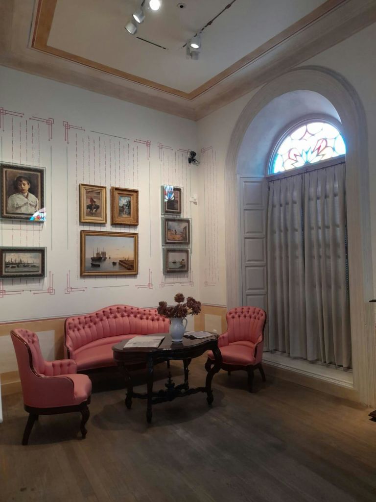 분홍색 빈티지 가구가 놓인 방, 벽에 액자가 놓인 그림, 꽃이 놓인 테이블, 커튼이 놓인 아치형 창문.