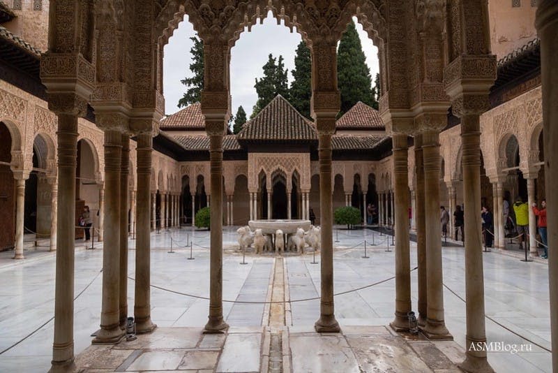 아치형 기둥, 사자 분수 센터피스, 정교한 조각, 주변의 타일 지붕이 있는 화려한 안뜰. 나무를 배경으로.