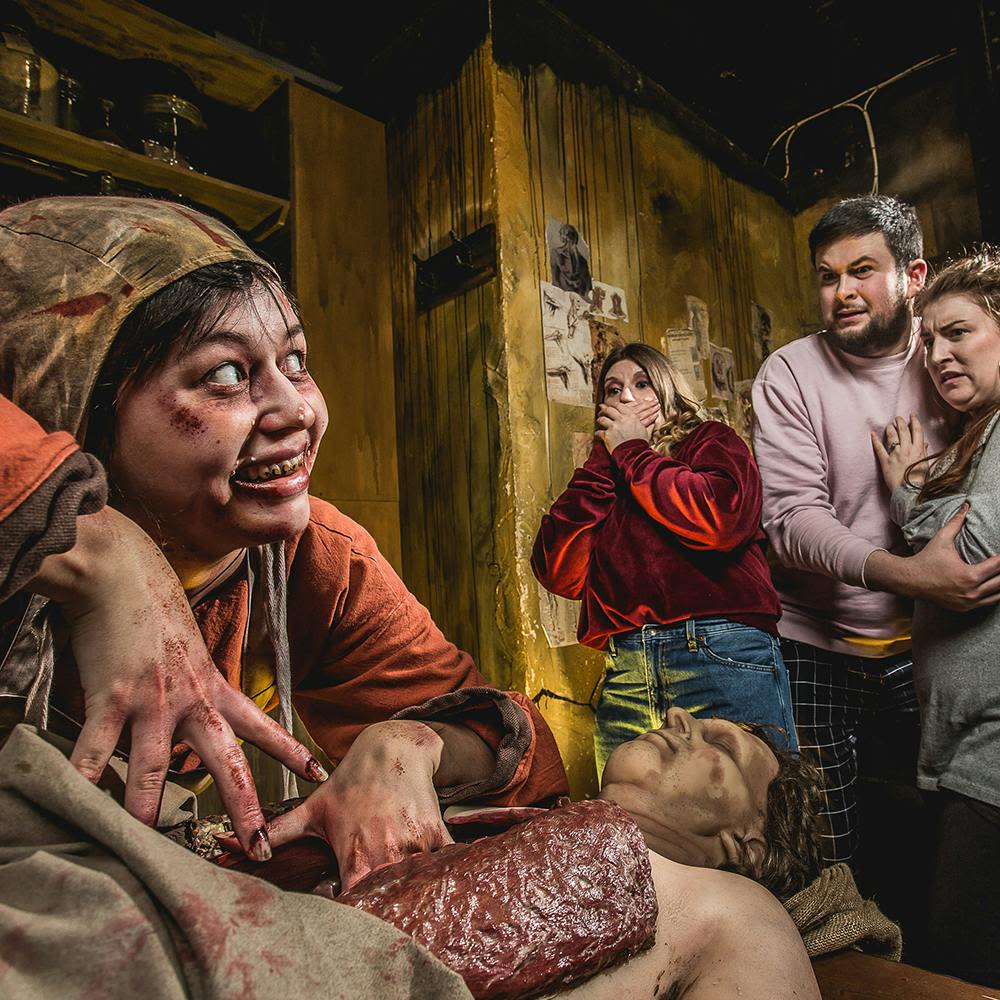 피투성이 손을 가진 사람이 조각난 마네킹 위로 위협적으로 웃으며 세 명의 구경꾼이 공포에 질려 반응합니다.