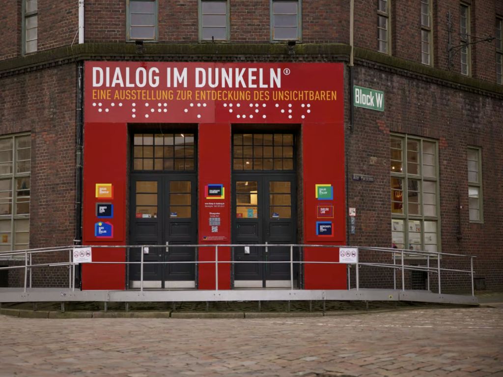 독일어로 된 표지판과 휠체어 램프가 있는 붉은 벽돌 건물의 "Dialog im Dunkeln" 전시관 입장.