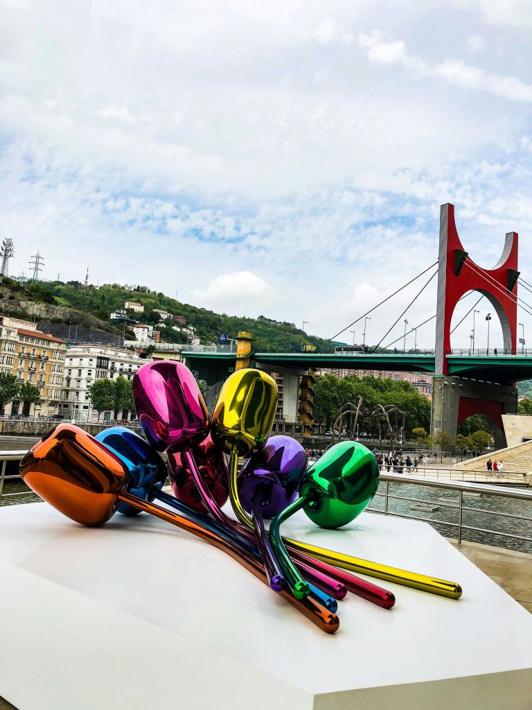 빨간색과 녹색의 다리, 건물, 언덕을 배경으로 한 받침대에 빛나는 화려한 풍선 조각품이 있습니다.