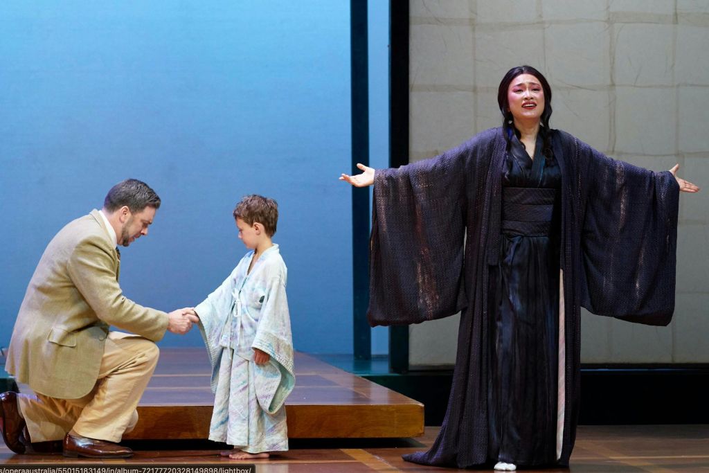 한 남자가 무릎을 꿇고, 한 아이가 가운을 입고, 한 여자가 어두운 가운을 입고 열정적으로 노래하는 연극 공연.