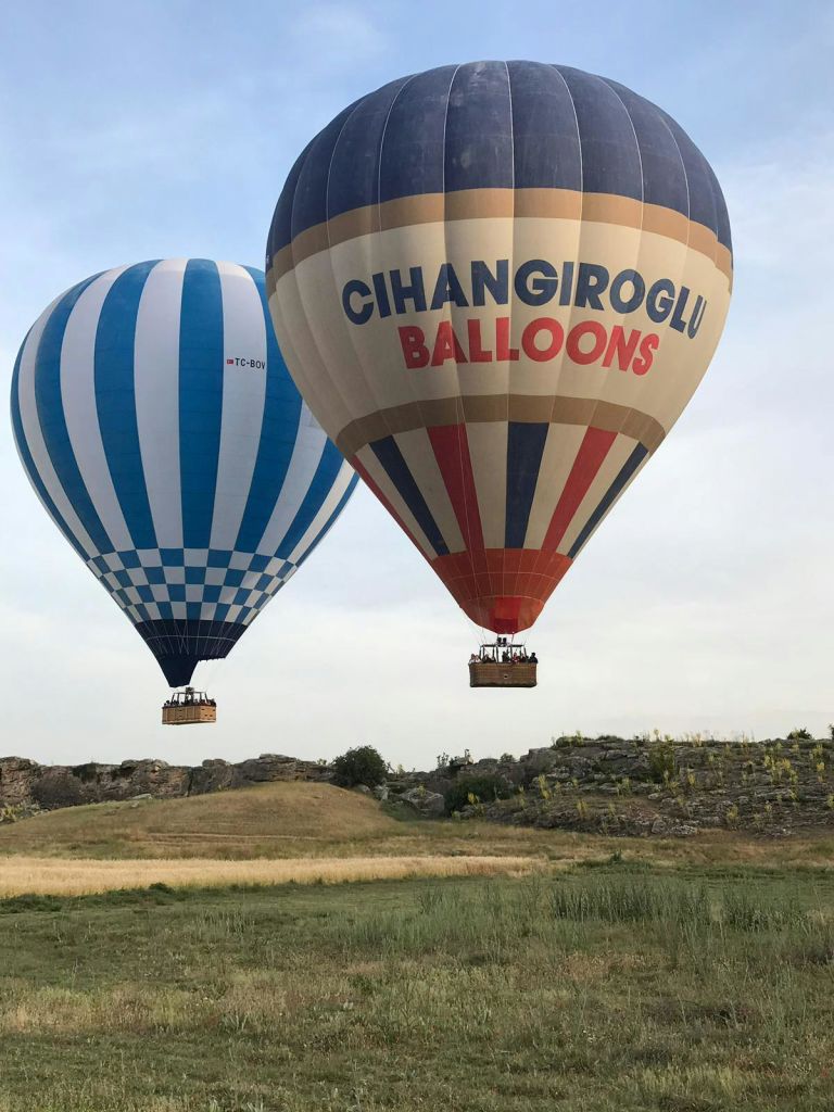파란 줄무늬가 있는 열기구 두 개와 "CIHANGIROGLU BALLOONS" 이라고 표시된 열기구 두 개가 잔디가 우거진 바위 지형 위로 날아오릅니다.